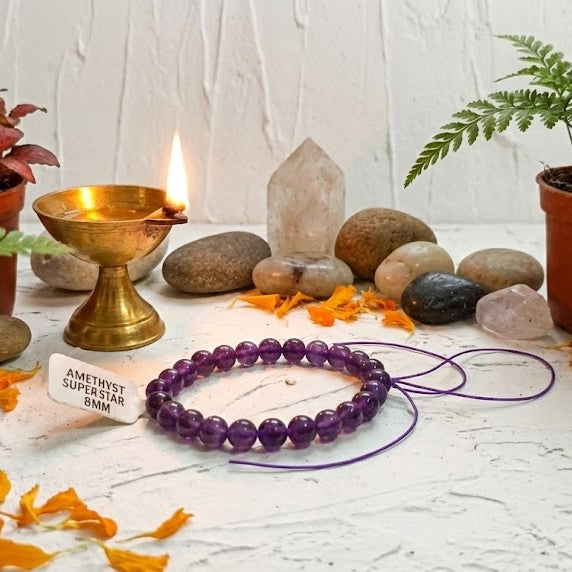 Decorative setting with purple beaded bracelet, candles, stones, and plants on a white surface.