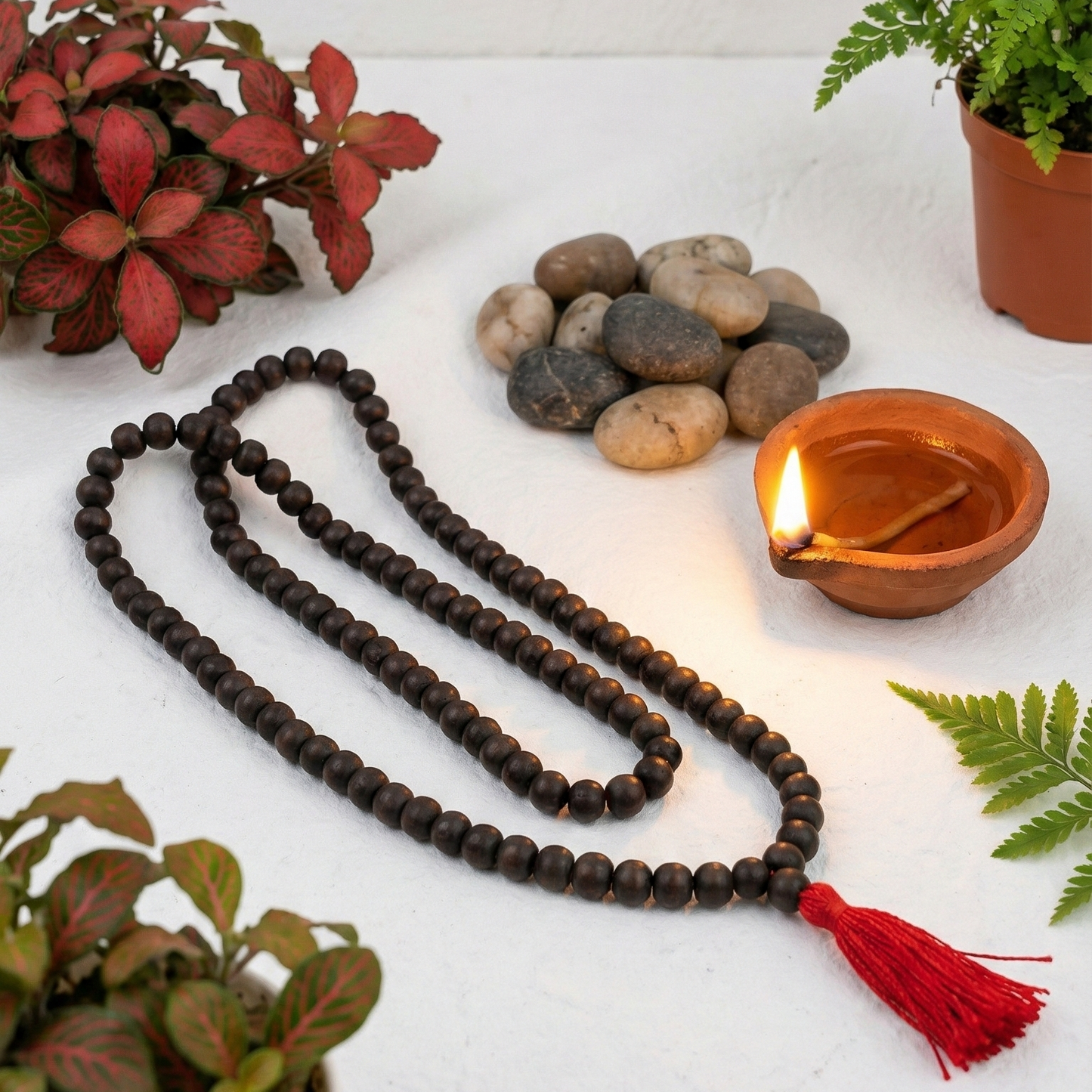 Meditation beads with a red tassel, rocks, and a lit candle on a white surface with plants.