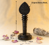 Black wooden incense stick holder with measurements and 'Original Ebony Wood' label, placed on a soft fabric background with dried flowers and a smudge stick.