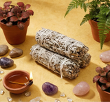 Two sage smudge sticks, a small bowl with a lighter, and crystals on a yellow surface.