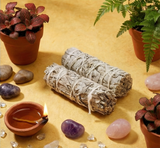 Two bundles of sage, a small bowl with a lit candle, crystals, and potted plants on a yellow surface.