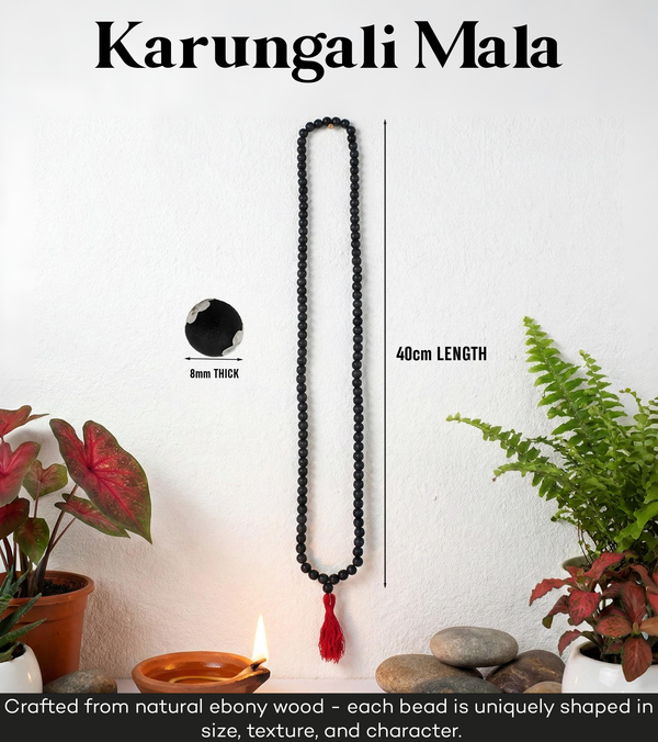 Black beaded mala necklace with a red tassel on a white background with plants and a candle.