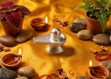 Silver Shiva Lingam surrounded by stones, candles, and plants on a yellow fabric background