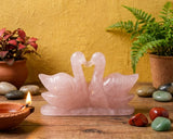 Rose quartz swan sculpture on a wooden surface with plants and stones.