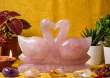 Rose quartz crystal swans on a yellow surface with plants and stones.