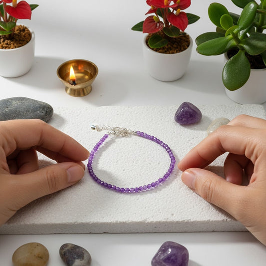 Purple beaded bracelet on a white surface with hands holding it, surrounded by small plants and a candle.