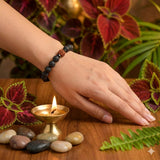 Hand wearing a beaded bracelet with a lit candle and pebbles on a wooden surface.