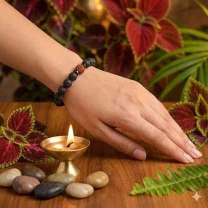 Hand wearing a beaded bracelet with a lit candle and pebbles on a wooden surface.