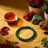 Green beaded bracelet on a textured surface with a lit candle, rocks, and plants.