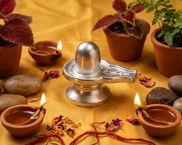 Silver Shiva lingam surrounded by potted plants, stones, and small oil lamps on a yellow fabric background.