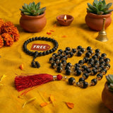 Black beaded mala with tassel on a yellow fabric background with marigold flowers and small plants.