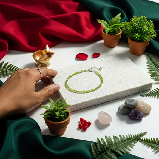 Hand holding a green bracelet on a white surface with plants, stones, and a lit oil lamp.