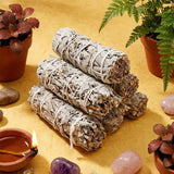 Stack of smudge sticks with a lit candle, crystals, and plants on a yellow surface